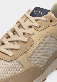 Μπεζ και καφέ αθλητικό παπούτσι με πάνελ από δίχτυ και σουέντ, επίπεδα κορδόνια, ραμμένες λεπτομέρειες και μπλε ετικέτα με την επιγραφή "CLAE EST. 2001" στη γλώσσα.