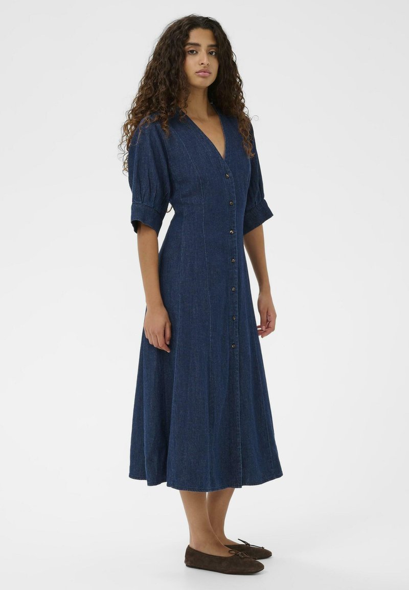 Jeans obleka z V-izrezom, kratkimi napihnjenimi rokavi, gumbom zadaj in tekočo A-linijo v temno modri barvi. V kombinaciji z rjavimi ravnimi čevlji.