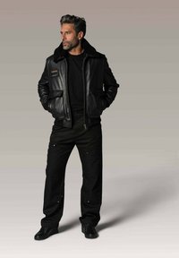 Homme en veste en cuir noir avec col en fourrure, chemise noire, pantalon noir et chaussures noires, se tenant avec les mains dans les poches en regardant à gauche.