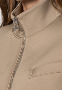 Chaqueta beige con cremallera y alto cuello, con cremallera metálica visible y un pequeño colgante en el tirador de la cremallera, llevada por una persona con cabello largo castaño.