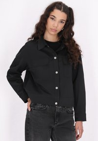 Sort cropped jakke med to brystlommer og sølvfarvede trykknapper. Har en glat tekstur og lange ærmer, parret med mørke jeans.
