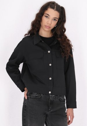 Sort cropped jakke med to brystlommer og sølvfarvede trykknapper. Har en glat tekstur og lange ærmer, parret med mørke jeans.