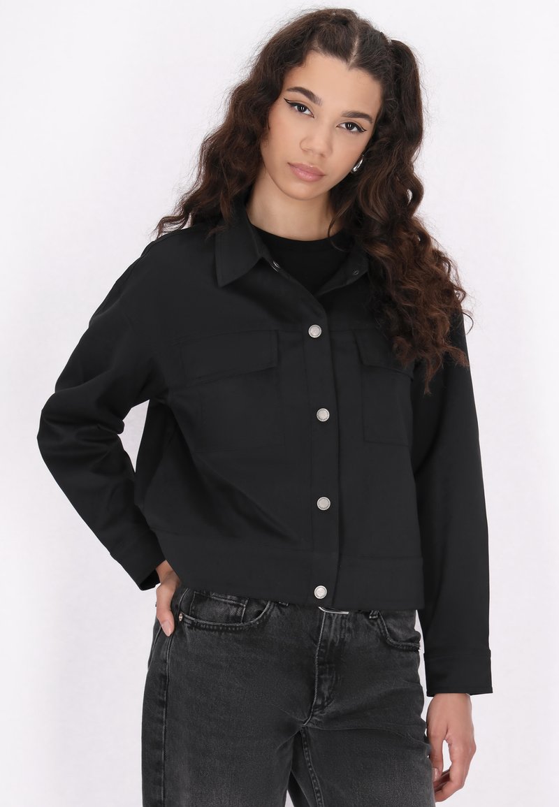 Sort cropped jakke med to brystlommer og sølvfarvede trykknapper. Har en glat tekstur og lange ærmer, parret med mørke jeans.