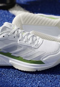 Scarpa atletica bianca con dettagli verdi, suola texturizzata e design laterale a strisce, posizionata su una superficie blu texturizzata.