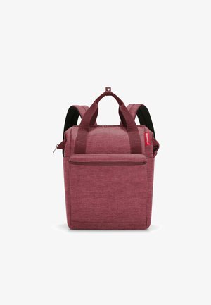 Mochila de tejido rojo con forma cuadrada, que cuenta con dos asas en la parte superior, correas de hombro acolchadas y un bolsillo frontal con cremallera. Superficie texturizada.