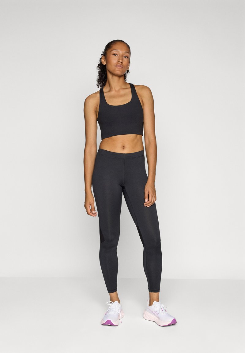 Schwarzes sportliches Cropped-Top und Leggings aus dehnbarem Material. Das Modell trägt pink-weiße Sneakers mit lila Akzenten. Einfaches Design.