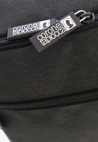 Bolsa de tela negra con una superficie lisa, cierre con cremallera y dos etiquetas distintas etiquetadas como "coronel taplacca".