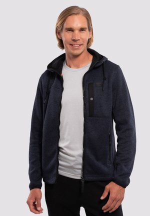Chaqueta de marina con cremallera, fabricada con tejido texturizado, detalles en negro, capucha y múltiples bolsillos con cremallera. Llevada sobre una camiseta gris claro.