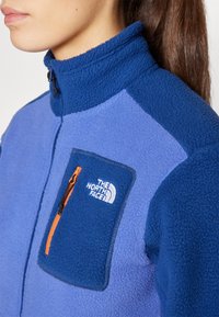 Polarowa kurtka w niebieskie i purpurowe panele, z wysokim kołnierzem, zapinana na zamek kieszeń na piersi z pomarańczowym zamkiem, z logo The North Face w białym kolorze.