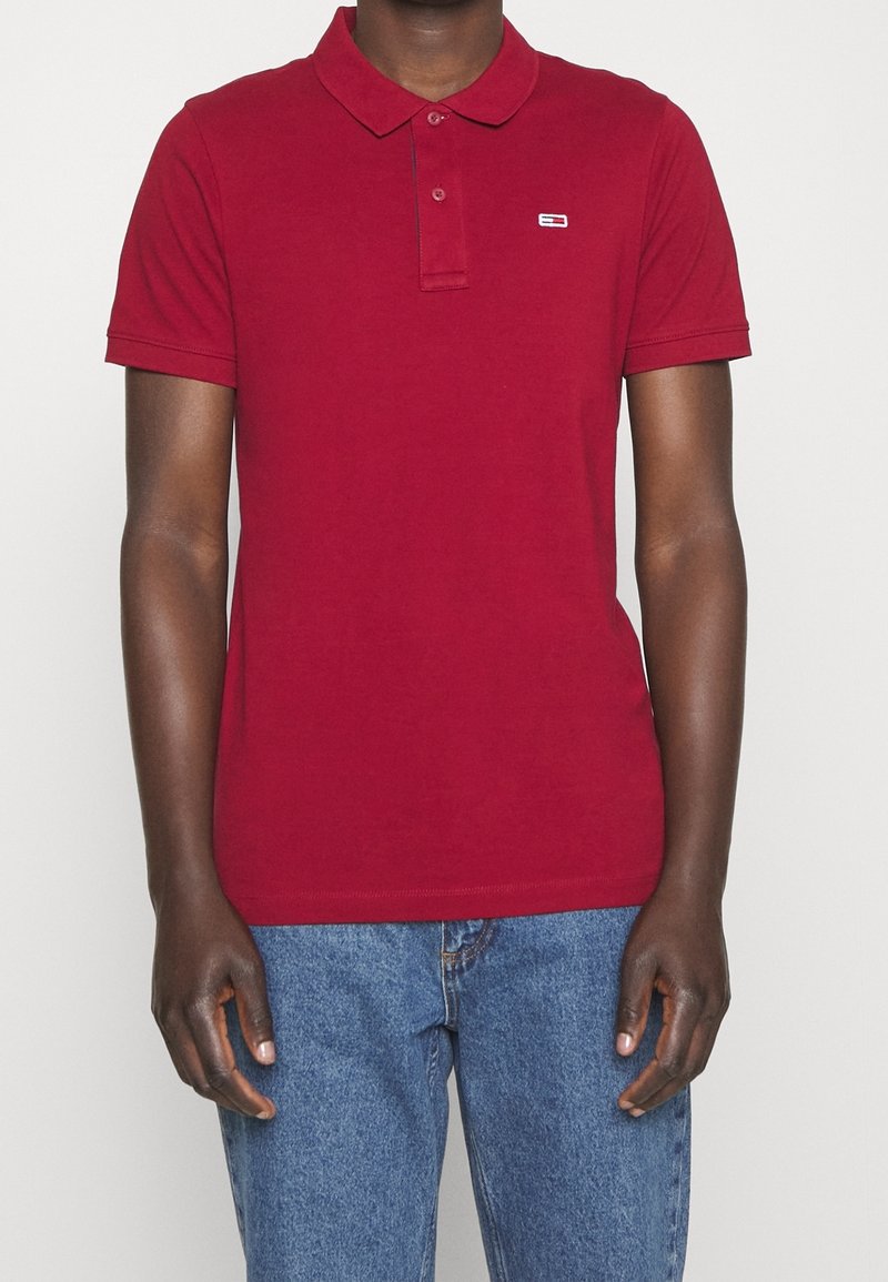 Polo rouge en coton, avec un col classique, une patte de boutonnage à deux boutons et un petit logo de marque sur la poitrine. Manches courtes, couleur unie.