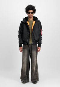 Jachetă bomber neagră cu căptușeală portocalie, pulover tricotat verde, blugi largi de culoare maro deschis și pantofi negri. Accesoriile includ ochelari de soare negri.