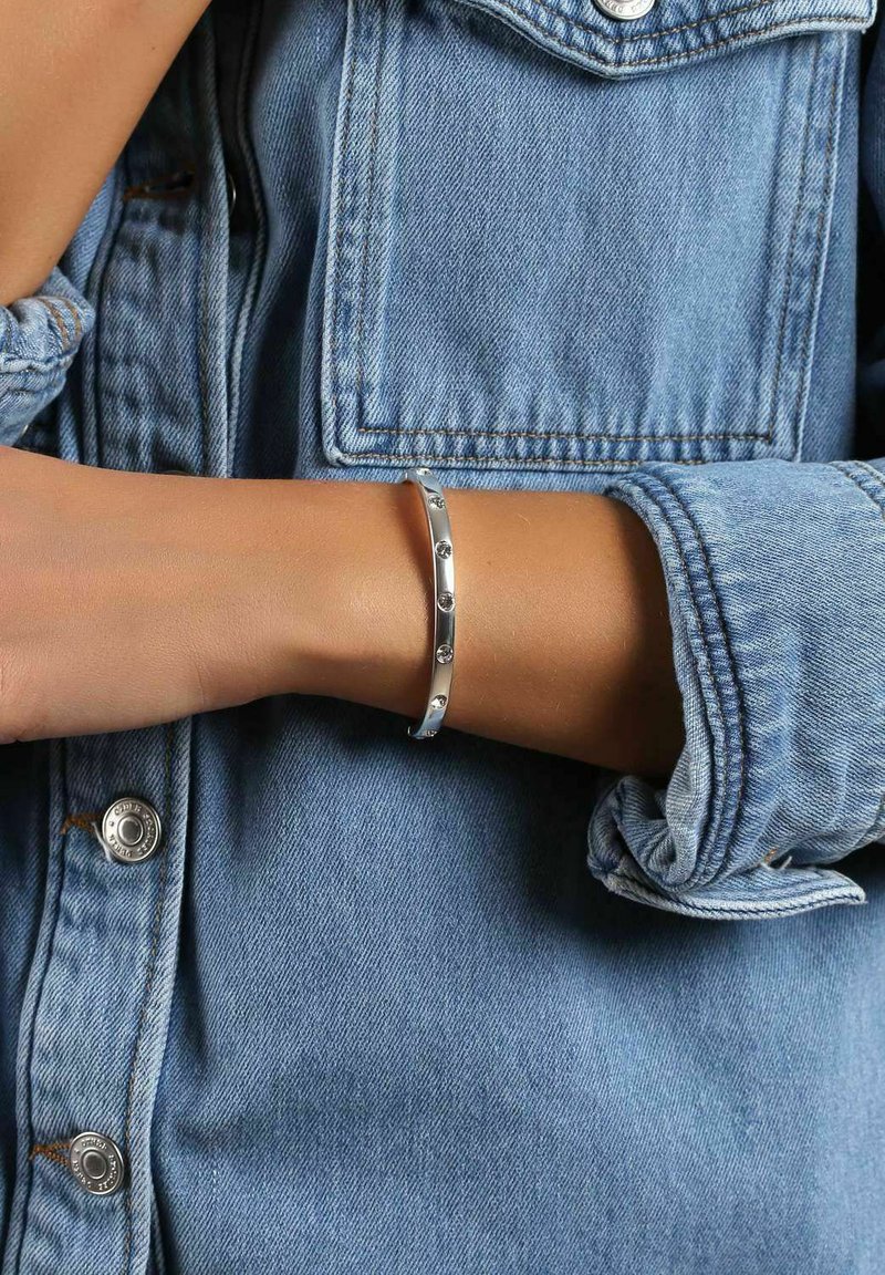 Silverarmband med cirkulära utskärningar och infällda ädelstenar, bärs på ett handleder mot en denimjacka med uppvikta ärmar.