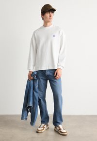 Camisa blanca de manga larga con pequeño logo azul, jeans de mezclilla azules, sosteniendo una chaqueta de mezclilla. Gorra marrón y zapatillas marrones y blancas.