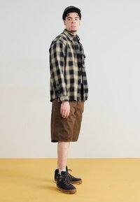 Cămașă flanelată cu carouri în negru și bej, pantaloni scurți maro, pantofi bleumarin cu tălpi maro. Modelul stă pe un fundal galben, împotriva unui fundal simplu.
