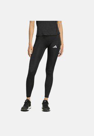 Donna che indossa leggings sportivi neri con il logo Adidas bianco sulla coscia e scarpe da ginnastica nere, in piedi contro uno sfondo uniforme.