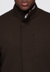 Hnědý kabát s texturou a vysokým límcem, vybavený předním zipem a nenápadnou boční kapsou. Čisté linie zdůrazňují jeho moderní design.