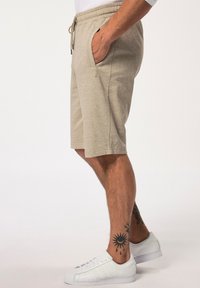 Shorts en tricot beige avec une taille élastique et une cordon de serrage, poches latérales, associés à des chaussures de sport blanches. Présente des tatouages sur les jambes.