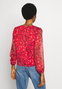 Rød blomstret bluse med gennemsigtige lange ærmer, rund halsudskæring og samlet manchet. Har et delikat mønster af hvide og blå blomster.