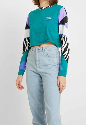 Persona con top corto de manga larga Vans en color azul verdoso con mangas de cebra y bloques de color, combinado con vaqueros de tiro alto en azul claro.