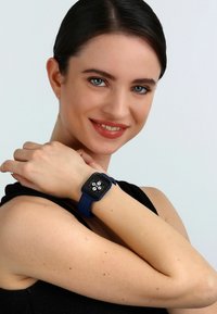 Smartwatch met een vierkante wijzerplaat, donker scherm en blauwe siliconen band. Toont vier ronde wijzerplaten op een zwarte achtergrond.