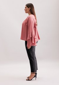 Top a maniche lunghe arricciato in morbido tessuto rosa chiaro, abbinato a jeans grigi scuri e sandali neri con tacco alto. Silhouette semplice e fluida.