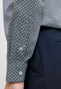 Cămașă cu mânecă lungă, prezentând un model geometric în albastru și alb, realizată dintr-un material texturat. Detaliu cu manșetă cu nasturi vizibil.