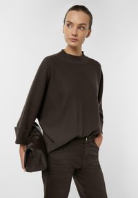 Brązowy sweter z dzianiny z wysokim kołnierzem i oversize'owymi rękawami, w połączeniu z ciemnobrązowymi dopasowanymi jeansami oraz brązową skórzaną torbą.