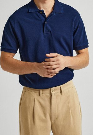 Polo bleu marine avec col, deux boutons et manches courtes, assorti à un pantalon plissé beige clair. Texture lisse et design décontracté.