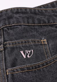 Μαύρο υφασμάτινο τζιν με πίσω τσέπη που έχει ροζ κεντημένο λογότυπο "V" και κίτρινα ραφές κατά μήκος των άκρων.