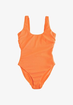 Mujer con bañador naranja apoyada en un poste de madera en una playa de arena con el océano y cielo azul al fondo.