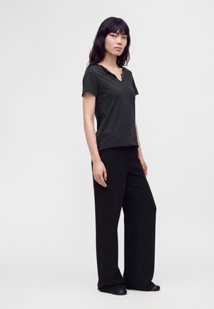 Femme aux longs cheveux noirs portant un haut noir à manches courtes boutonné et un pantalon noir à jambes larges, debout devant un fond clair uni.