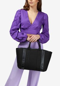 Bolso tote negro con detalles de texto en relieve, asas resistentes y una superficie texturizada. Combinado con una blusa morada y pantalones morados claros.