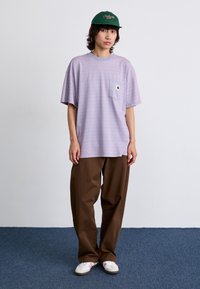 T-shirt rayé violet avec une poche poitrine, associée à un pantalon marron ample et des baskets blanches. Le mannequin porte une casquette verte avec un texte rouge.