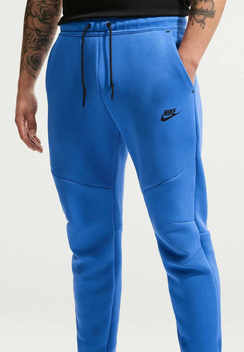 Personne portant un pantalon de survêtement Nike bleu vif avec des cordons noirs, des tatouages visibles sur les bras et une main dans la poche.