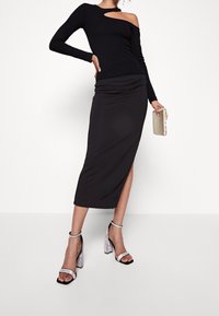 Frau trägt ein schwarzes asymmetrisches Langarmoberteil, einen schwarzen Midirock mit seitlichem Schlitz, silberne High-Heels und hält eine beige Clutch.