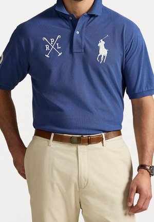 Man wearing blue polo shirt with white crossed golf clubs and riding polo player logos, beige pants, brown belt, and wristwatch.