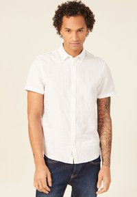 Camicia bianca in lino a maniche corte con colletto a bottone e maniche arrotolate, abbinata a jeans in denim blu. Presenta una vestibilità casual.