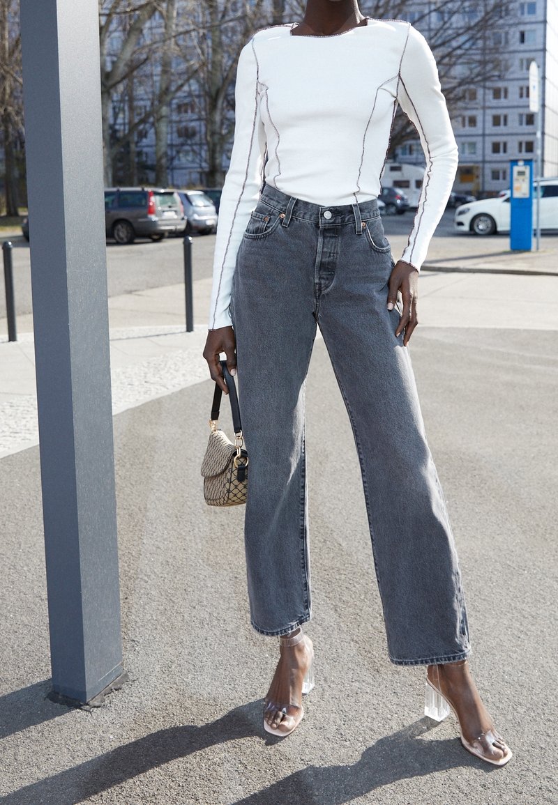 Femme portant un haut blanc à manches longues avec coutures noires, jean gris taille haute, sandales à talons transparents, tenant un petit sac à main à motifs.