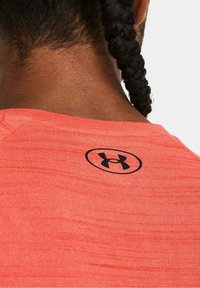 Světlé korálové sportovní tričko s texturovaným povrchem, s černým logem Under Armour na horní části zad. Standardní design s kulatým výstřihem.