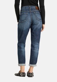 Jean en denim bleu foncé à taille haute, revers roulés, cinq poches, et une étiquette en cuir à l'arrière. Texture lisse avec des détails d'usure.