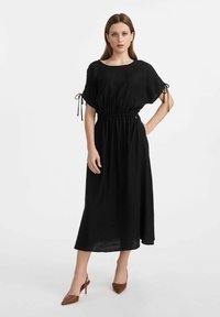Czarna midi sukienka o luźnym fasonie, elastycznym pasie, krótkich rękawach z wiązaniami i zwiewnej sylwetce. Noszona z brązowymi czółenkami z ostrym noskiem.
