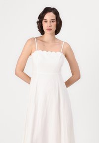 Abito bianco con scollatura a onde, spalline sottili e silhouette fluttuante realizzato in tessuto leggero, caratterizzato da una consistenza liscia.