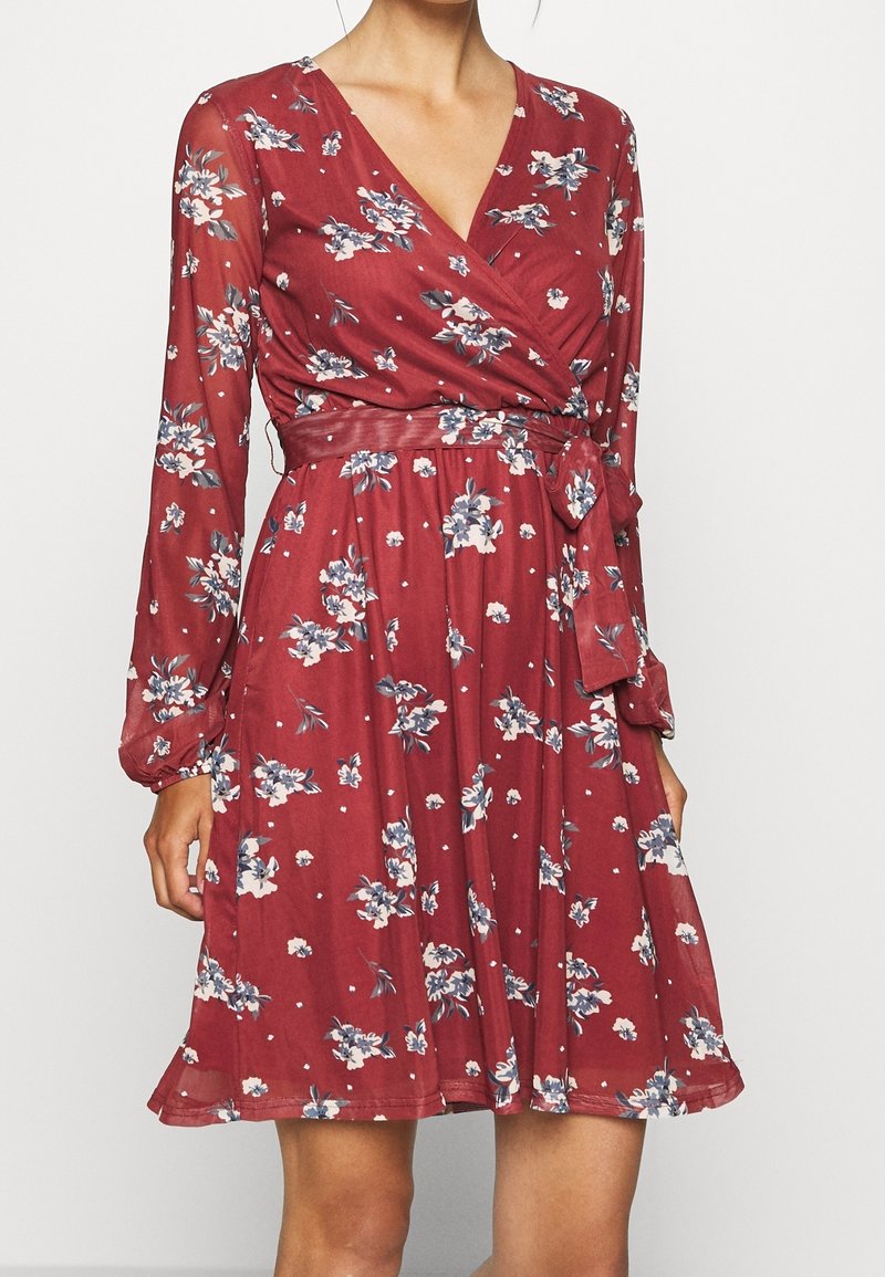 Femme portant une robe portefeuille rouge à motifs floraux, manches longues et ceinture nouée à la taille, debout devant un fond blanc.