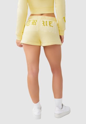 Femme portant un short jaune pâle avec le texte doré pailleté "TRUE" au dos, un sweat-shirt court assorti, des chaussettes blanches et des baskets blanches.