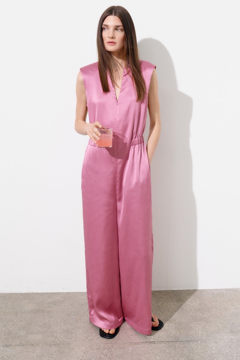 Femme aux longs cheveux bruns portant une combinaison rose en satin sans manches, tenant un verre de boisson rose, debout contre un mur blanc.