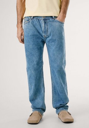 Homme portant un jean bleu clair ample, des chaussures beiges sans lacets et une chemise jaune clair rentrée, debout avec une main dans la poche.