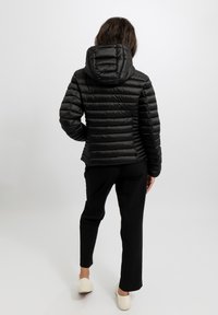 Giacca imbottita nera con cuciture orizzontali, cappuccio e polsini elastici. Indossata con pantaloni neri e scarpe bianche, vista da dietro.