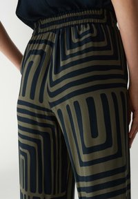 Hooggetailleerde broek van olijfgroene stof met een donkerblauwe geometrische patroon. Beschikt over een elastische tailleband en een gladde textuur.