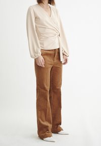 Top avvolgente beige a maniche lunghe con un dettaglio a cravatta, abbinato a pantaloni a zampa in corduroy marroni e scarpe bianche a punta. Tessuti lisci e textured.