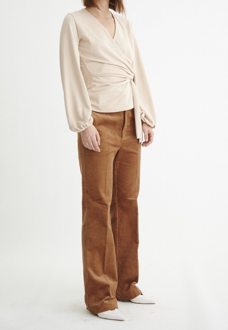 Top avvolgente beige a maniche lunghe con un dettaglio a cravatta, abbinato a pantaloni a zampa in corduroy marroni e scarpe bianche a punta. Tessuti lisci e textured.